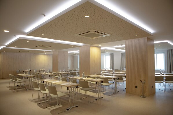 A conference room with multiple tables and chairs, large windows, and a high ceiling with recessed lighting.