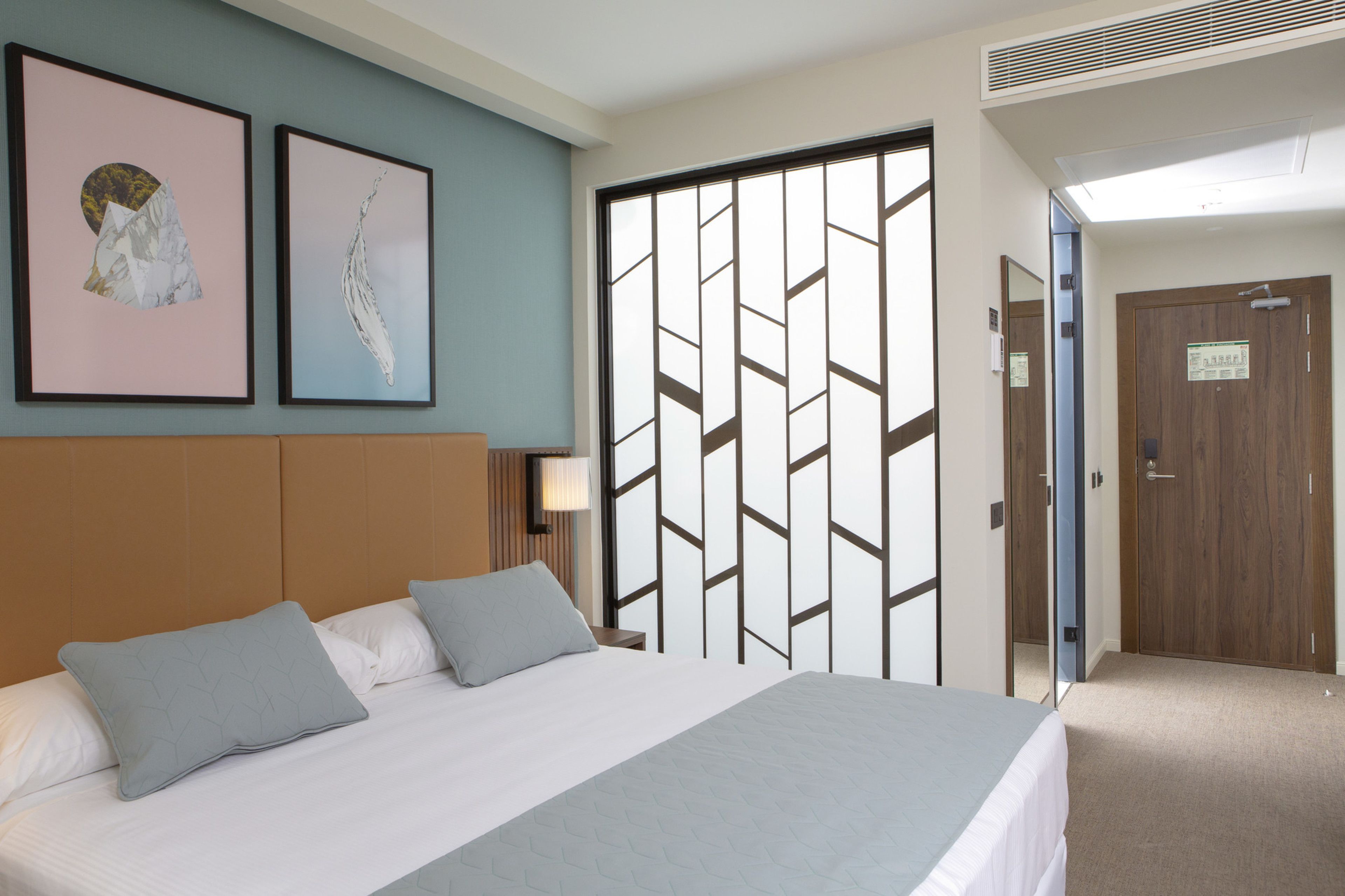 Une chambre d’hôtel avec un lit, deux oreillers, une tête de lit, une lampe fixée au mur et deux tableaux encadrés.