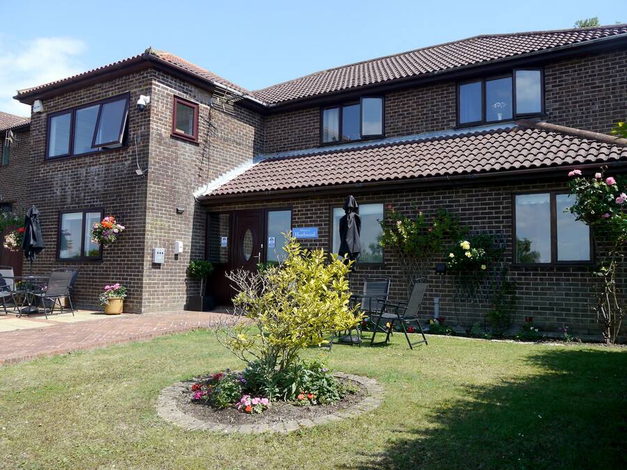 A two-story brick building with a tiled roof, a front door, and a small garden area with a tree and flower beds.