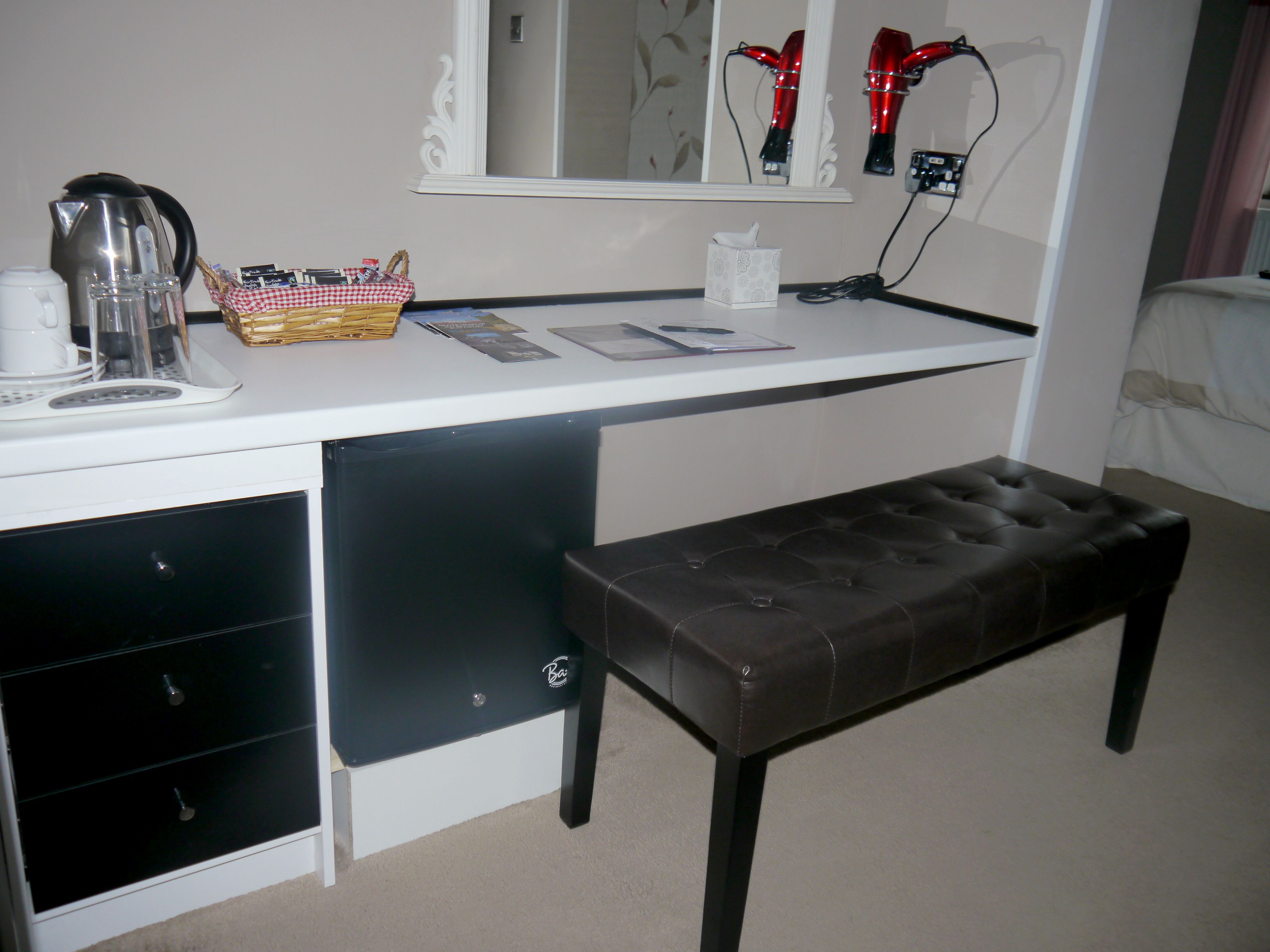 A hotel room with a white desk, a black bench, a mirror, and a bed.