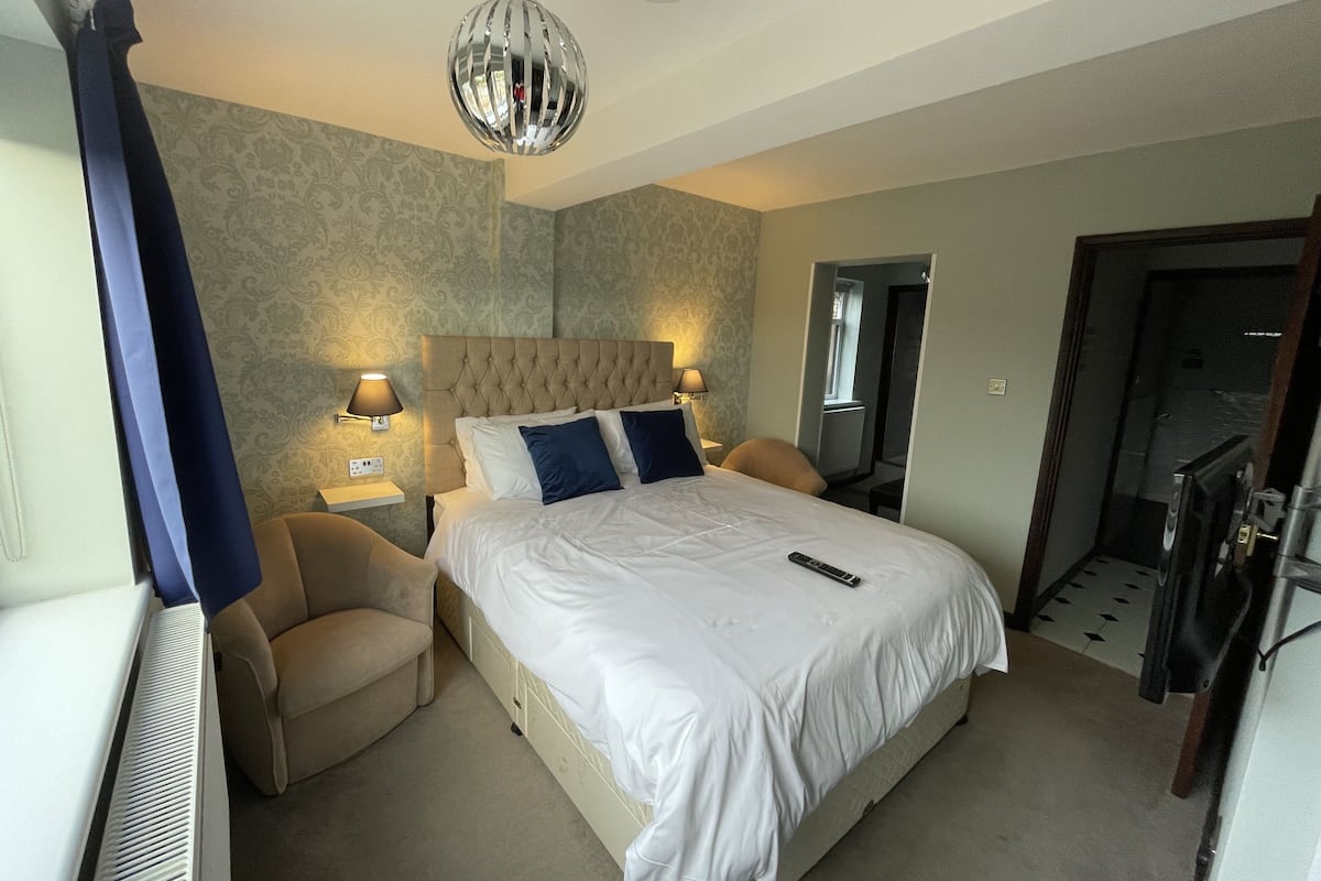 A hotel room with a large bed, two bedside lamps, a ceiling light, a radiator, and a door leading to another room.