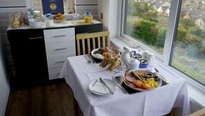 A breakfast table with a variety of food items, a view of a residential area through a window, and a small cabinet with kitchen items. - Heathwood BnB (Dover)