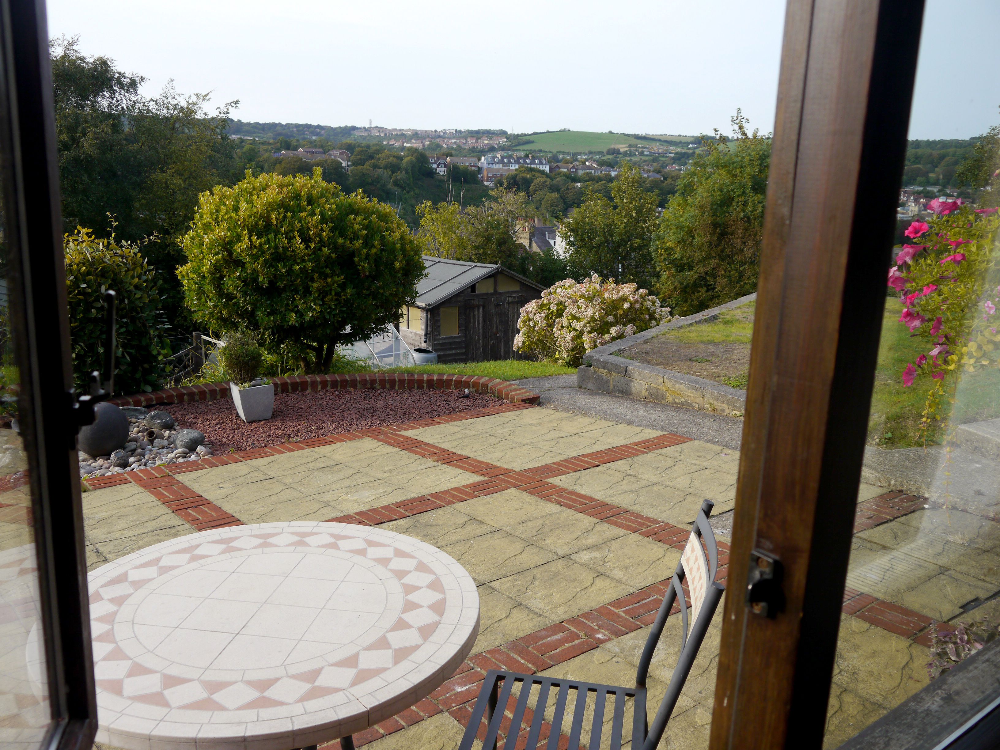 A patio with a tiled table and chairs, surrounded by a brick border and a garden with trees and flowers.