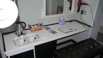 Et hotelværelse med et hvidt skrivebord, en sort bænk, et spejl, en hårtørrer, en kurv, en tissueskasse, en elkedel, en kop og nogle papirer.