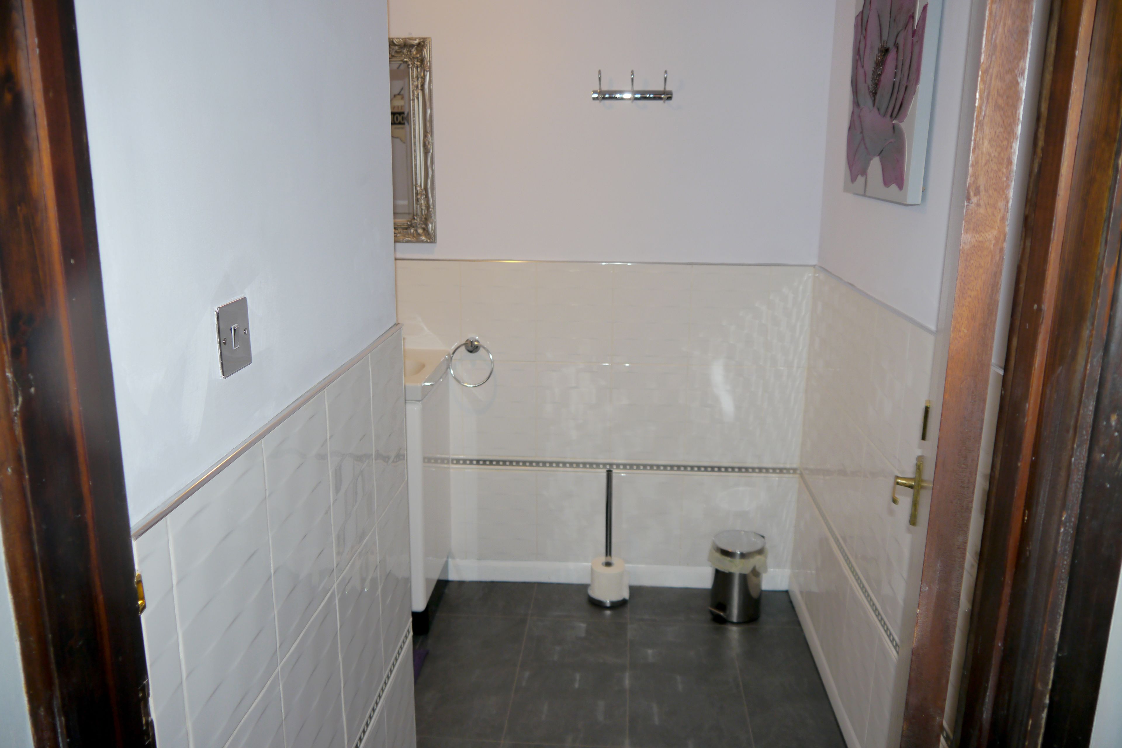 A bathroom with white tiled walls, a dark floor, a toilet, a trash bin, a towel rack, and a mirror.