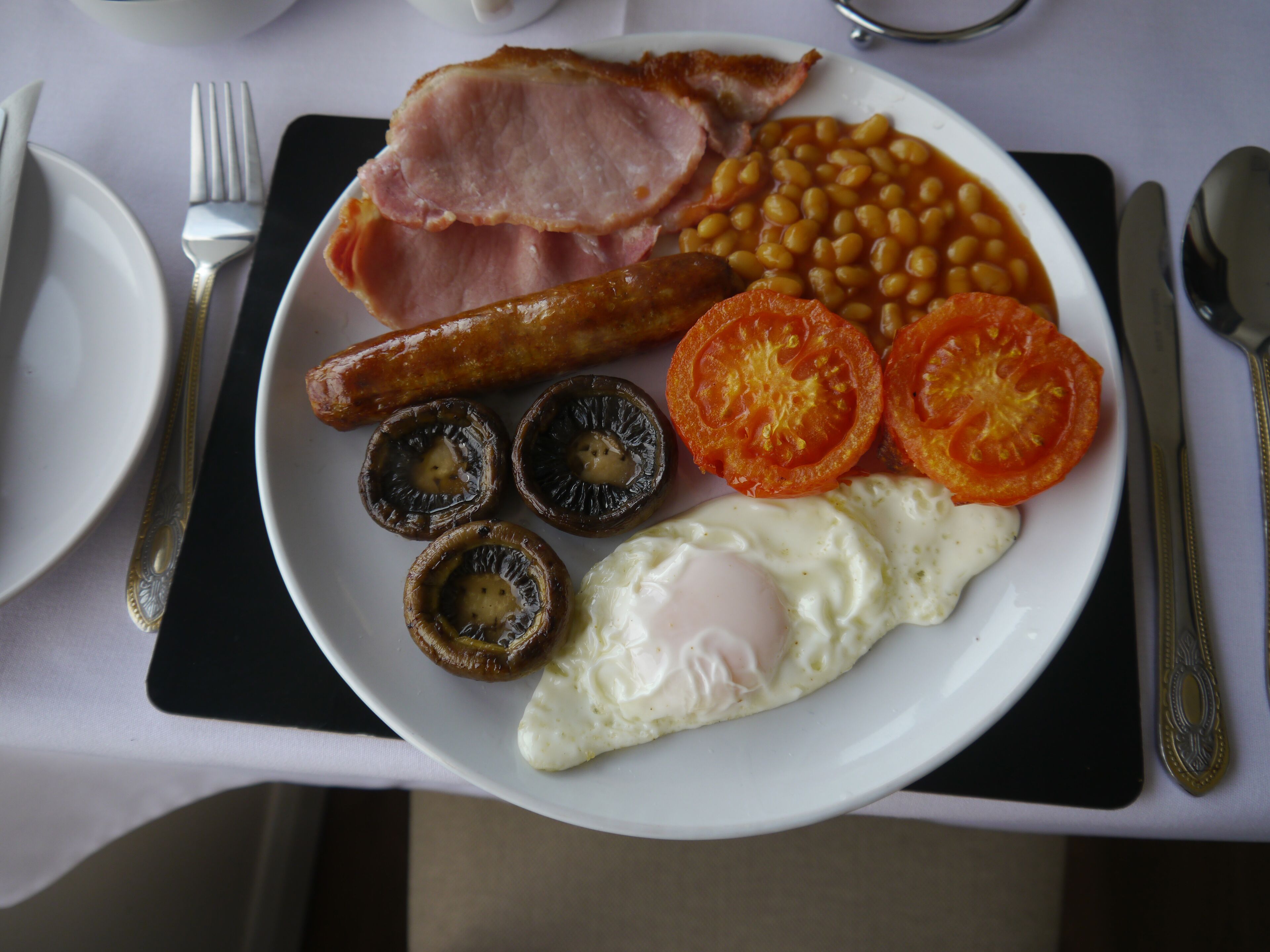 A breakfast plate with eggs, bacon, sausages, baked beans, tomatoes, and mushrooms.