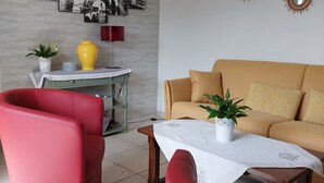Un salon comprenant un canapé, deux fauteuils rouges, une table basse et une petite table avec une lampe. Les murs sont décorés, notamment avec une représentation d’un paysage urbain et des miroirs ronds.