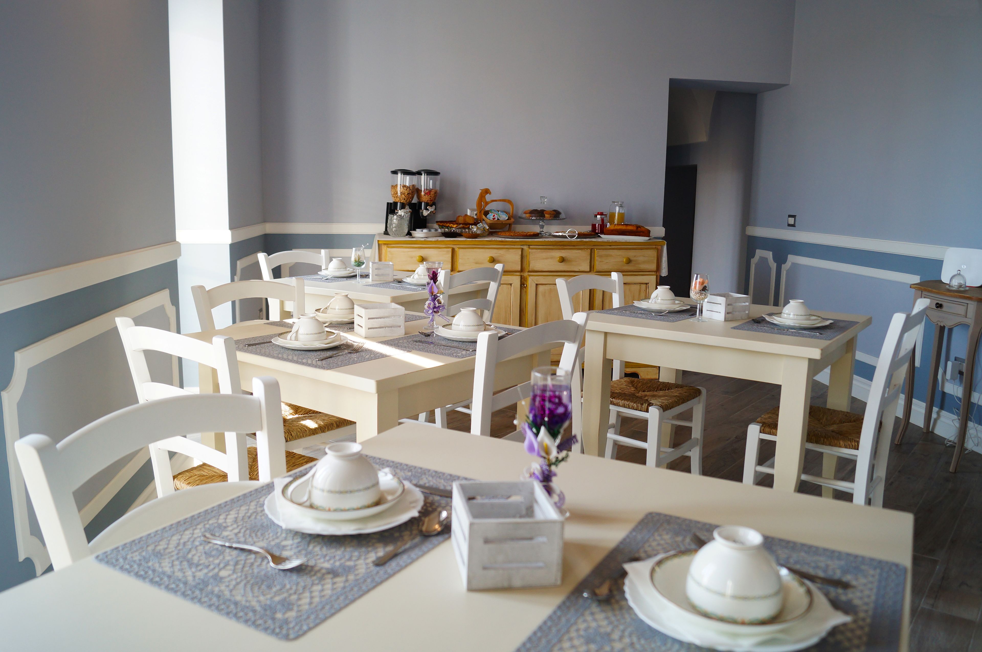 A dining area with white chairs and tables, a wooden buffet counter, and a vase with purple flowers.