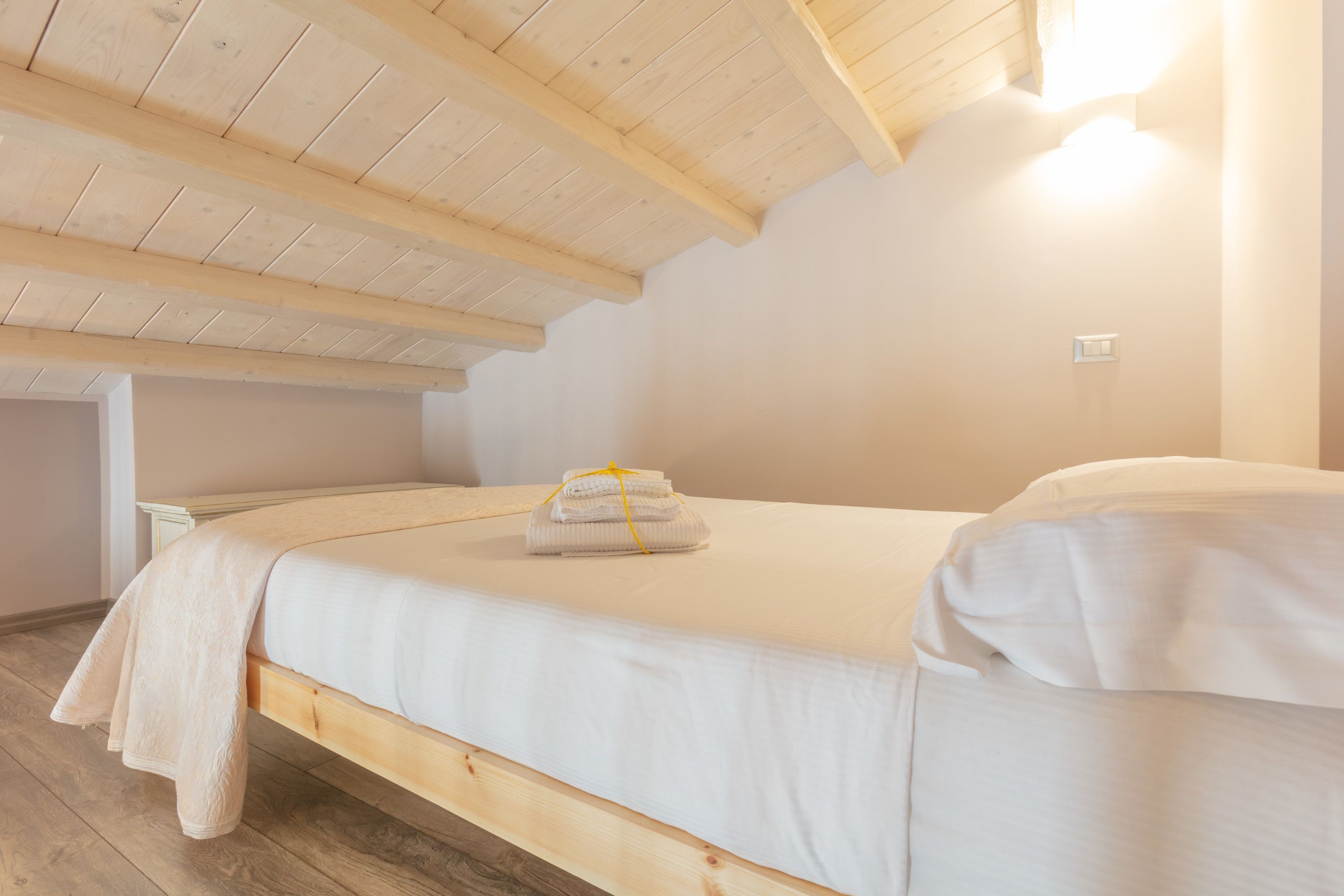 A single bed in a room with wooden beams on the ceiling, a white wall, and a light switch.