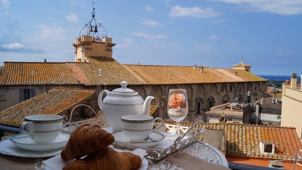 Eine Terrasse mit Tisch, gedeckt für Tee, einem Croissant und einem Weinglas, mit Blick auf eine Stadtlandschaft mit Ziegeldächern und historischen Gebäuden.