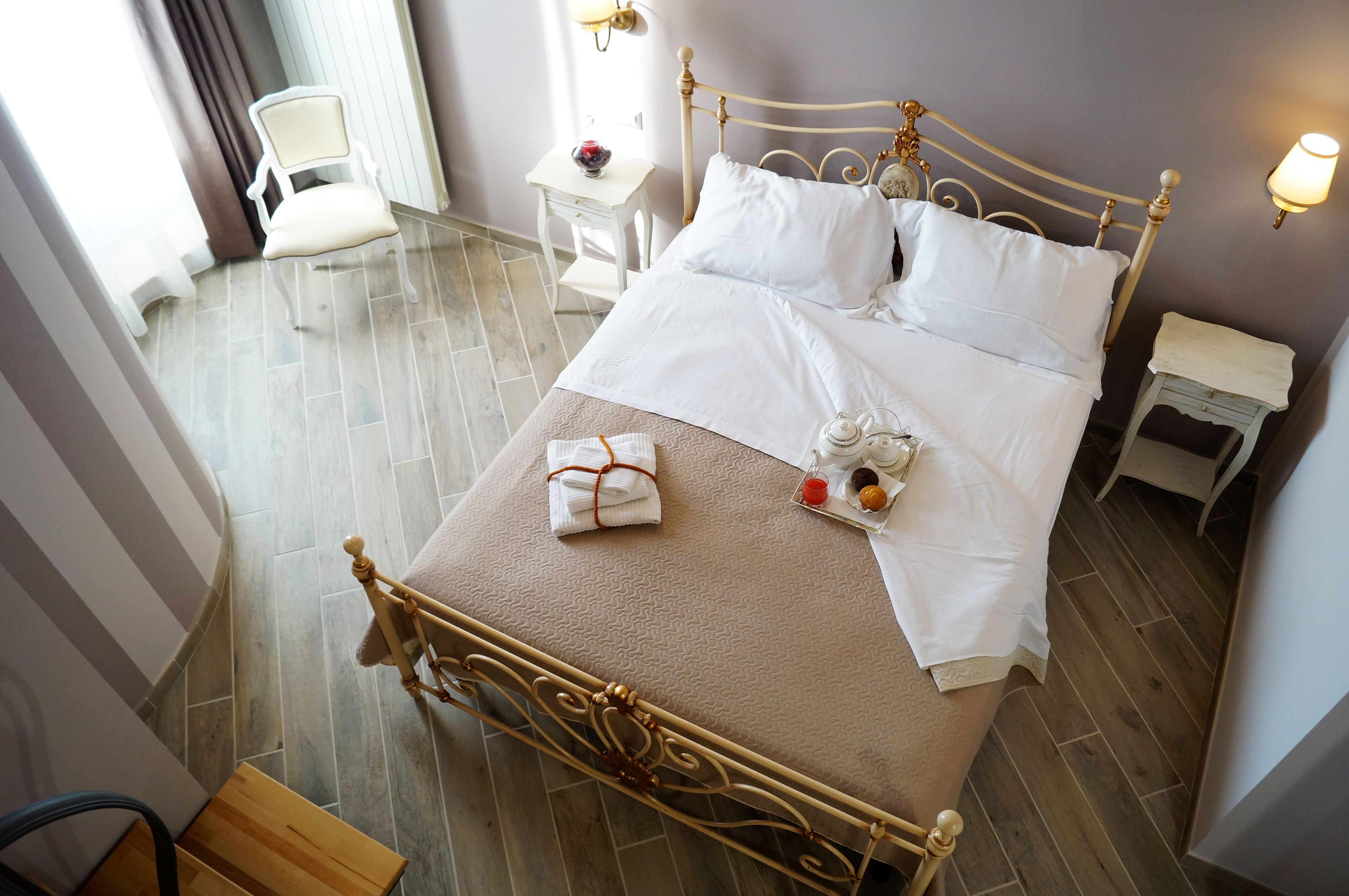 Uma cama bem arrumada com roupa de cama branca, uma mesinha de cabeceira com abajur e uma cadeira em um quarto com piso de madeira e parede curva.