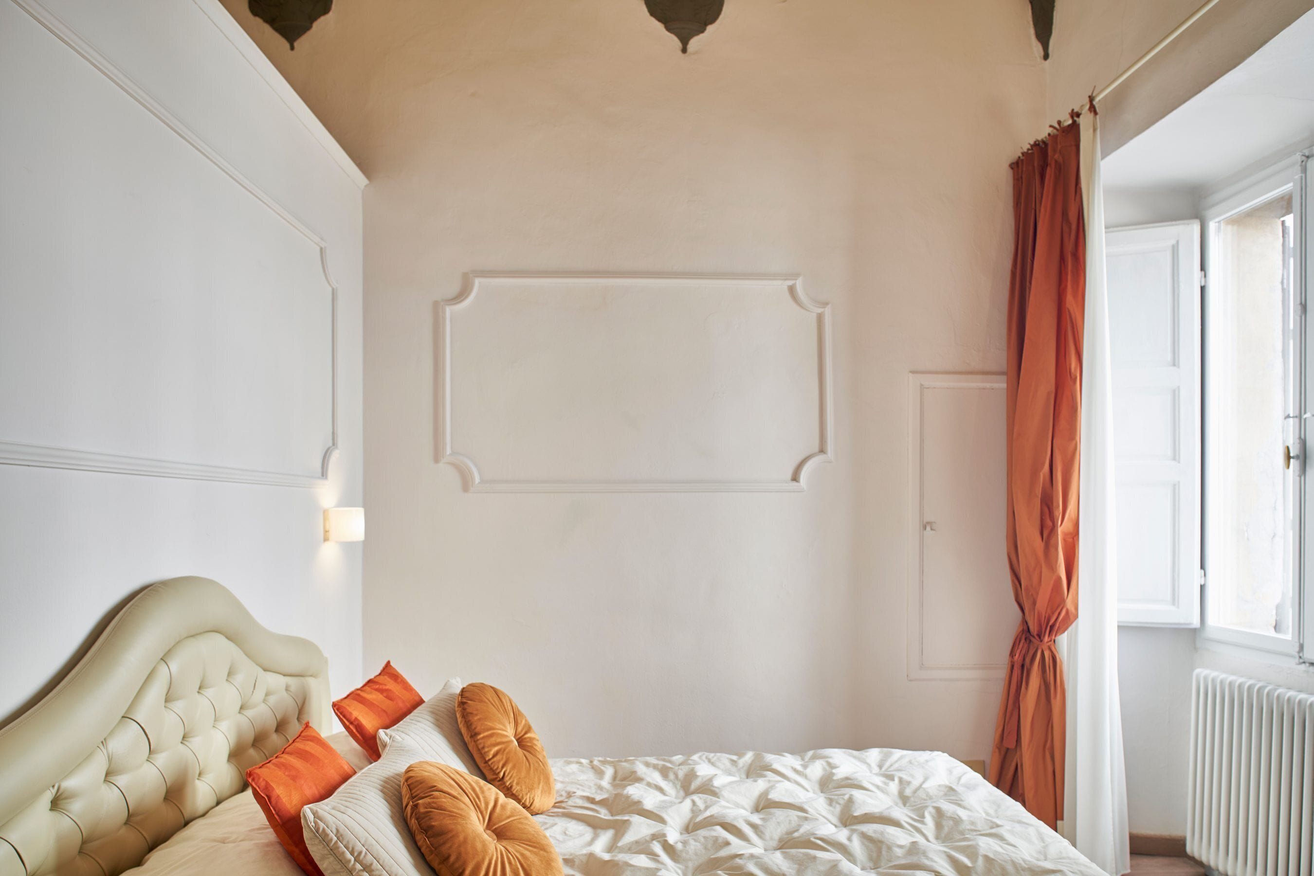 A bedroom with a tufted headboard, a large bed with white bedding, two orange pillows, and a window with an orange curtain.