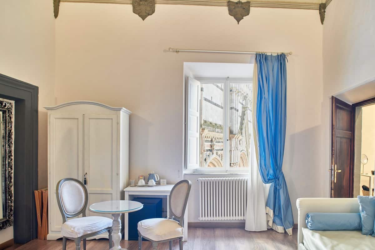 A room with a white cabinet, two chairs, a small table, a radiator, a window with blue curtains, and a white sofa with blue cushions.