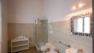 A bathroom with a glass-enclosed shower, a toilet, a bidet, a vanity with a mirror, and a shelf with towels.