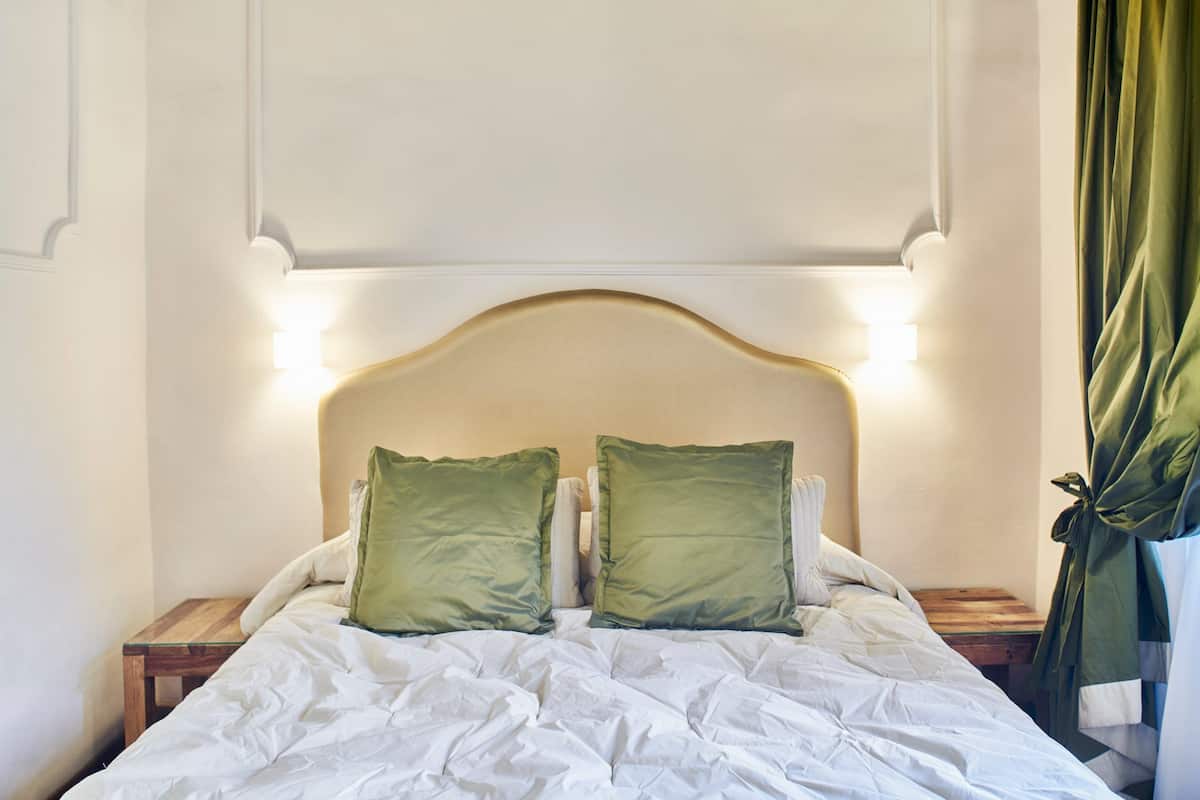 A neatly made bed with white linens and two green pillows, a wooden nightstand beside it, and a green curtain on the window.