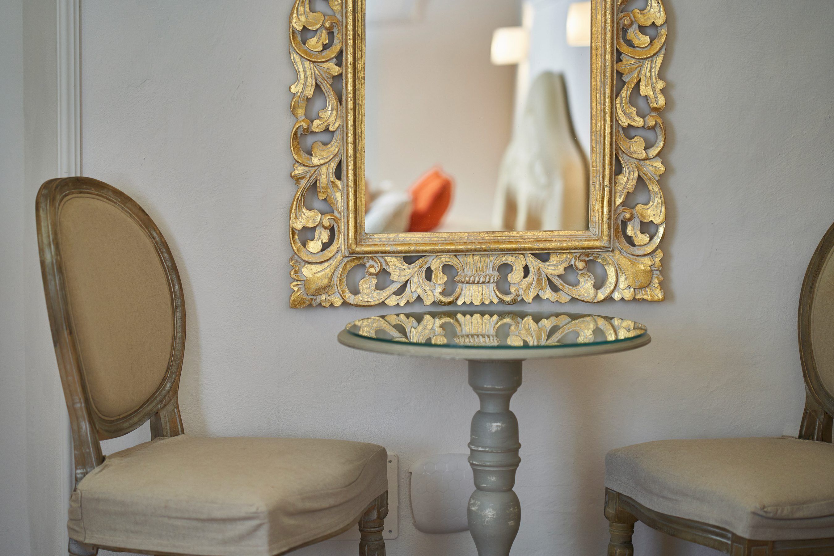 Due poltrone antiche con sedute e schienali imbottiti, poste su ciascun lato di un piccolo tavolino rotondo con piano in vetro. Sopra il tavolo, uno specchio decorativo con cornice dorata finemente lavorata.