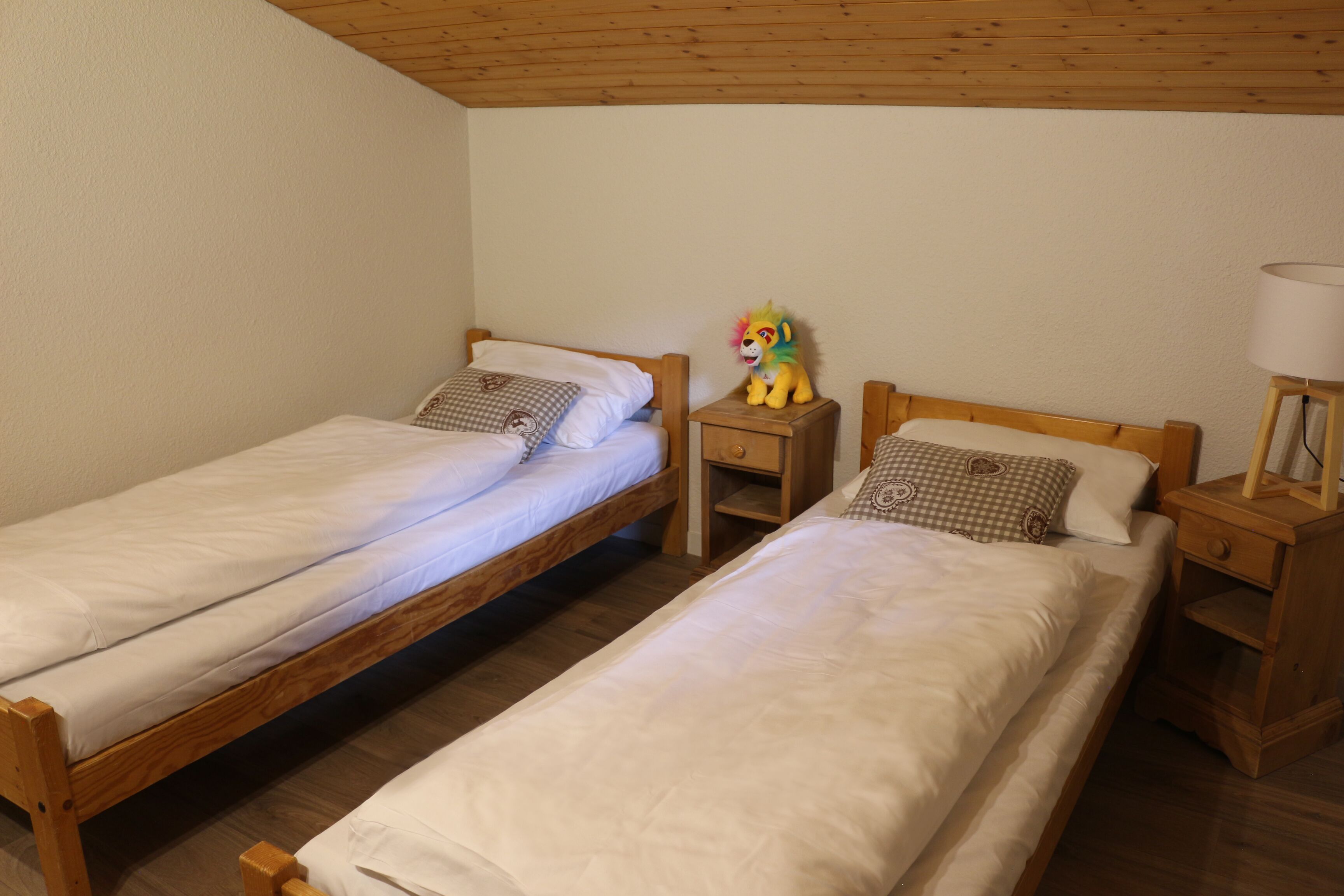 Een kamer met twee houten bedden, elk voorzien van wit beddengoed en decoratieve kussens. Aan één kant staat een klein houten nachtkastje met een lamp en een pluchen speelgoed.