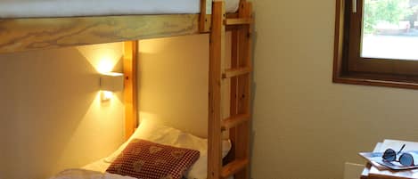 A bunk bed room with a wooden ladder, a small desk with a chair, and a window with a view of greenery.