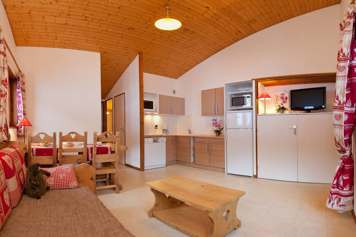 Une pièce chaleureuse avec un plafond en bois, une table en bois, un canapé, une petite chaise en bois, un lit recouvert d’une couverture à carreaux rouges et blancs, et un coin cuisine équipé d’un four à micro-ondes, d’un réfrigérateur et d’une télévision.