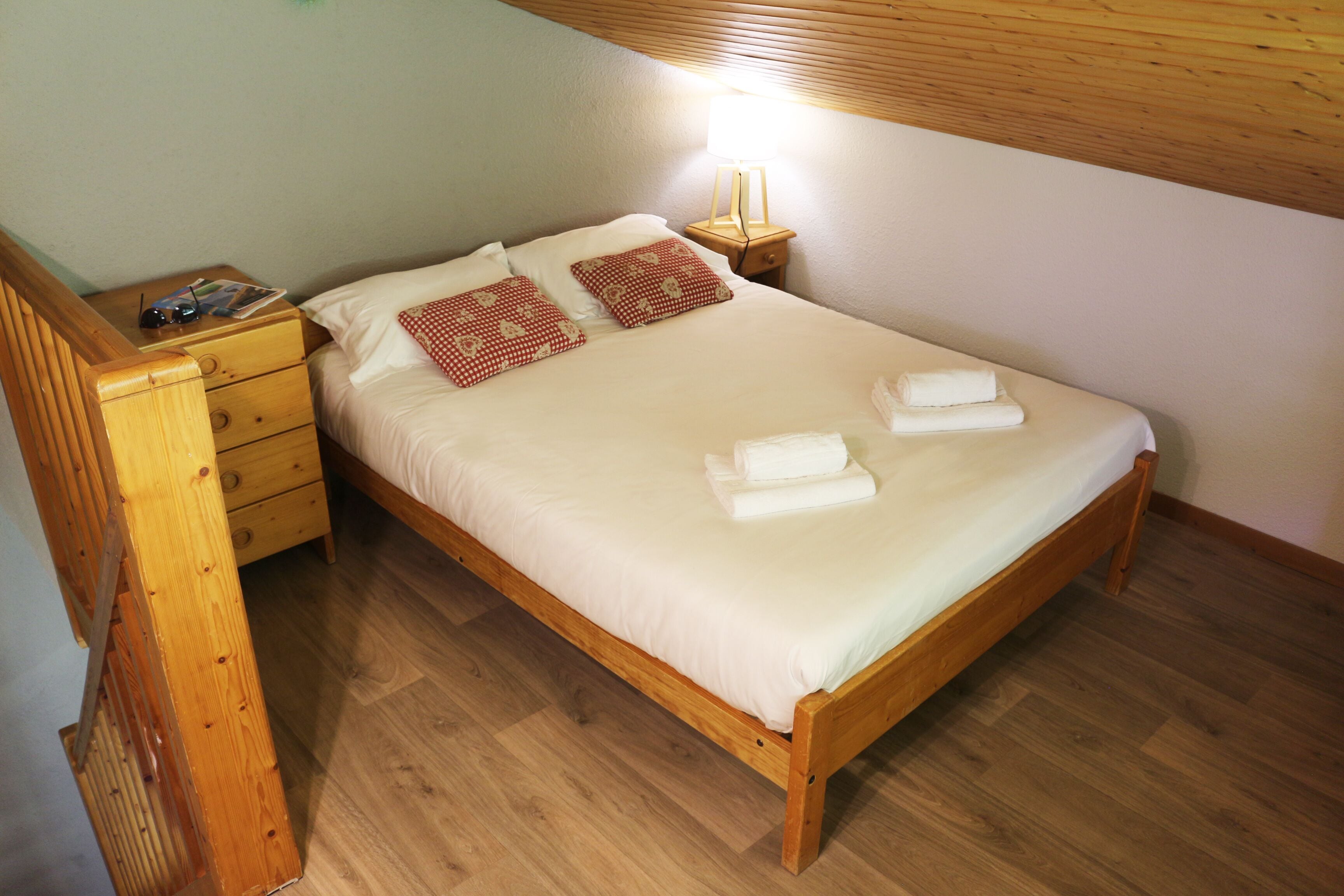 A neatly made bed with white linens and two red pillows, a wooden nightstand with a lamp, and a wooden dresser with drawers.