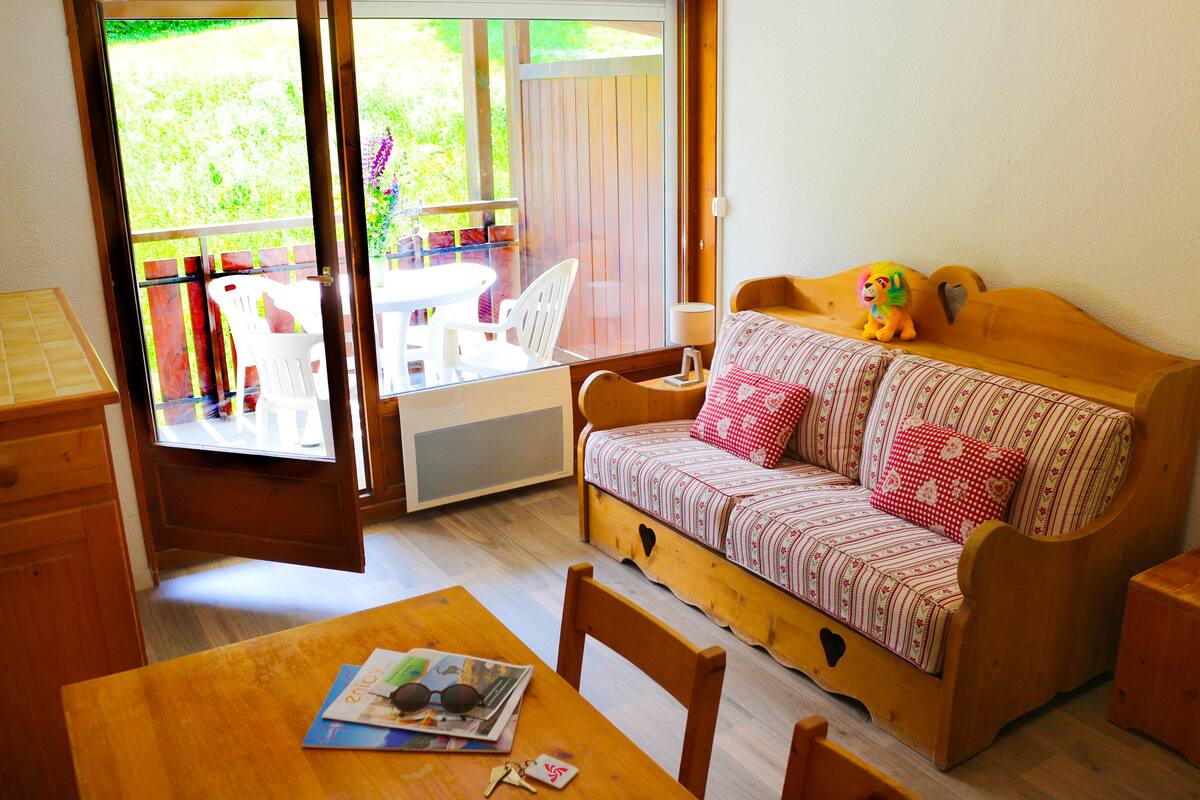 Une pièce chaleureuse avec un canapé en bois, une table à manger et une vue sur un jardin, visible à travers une porte-fenêtre coulissante.