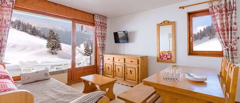 Una habitación acogedora con una cama de madera, una mesa de comedor con platos y cubiertos, un televisor en la pared y vistas a un paisaje nevado a través de las ventanas.