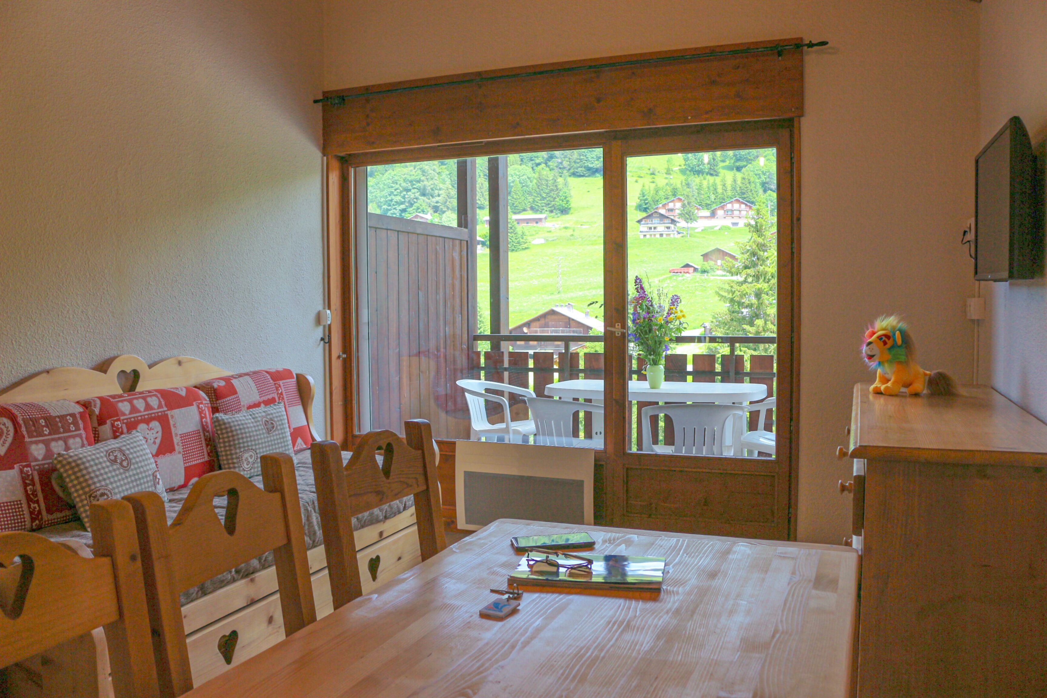 A room with a wooden table, chairs, a bed with a quilt, and a view of a garden through a sliding door.