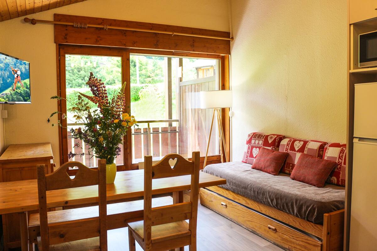 Une table de salle à manger en bois, accompagnée de chaises, un canapé avec des coussins à motifs, un vase rempli de fleurs et un tableau accroché au mur.