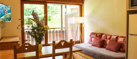 A wooden dining table with chairs, a sofa with patterned cushions, a vase with flowers, and a painting on the wall.