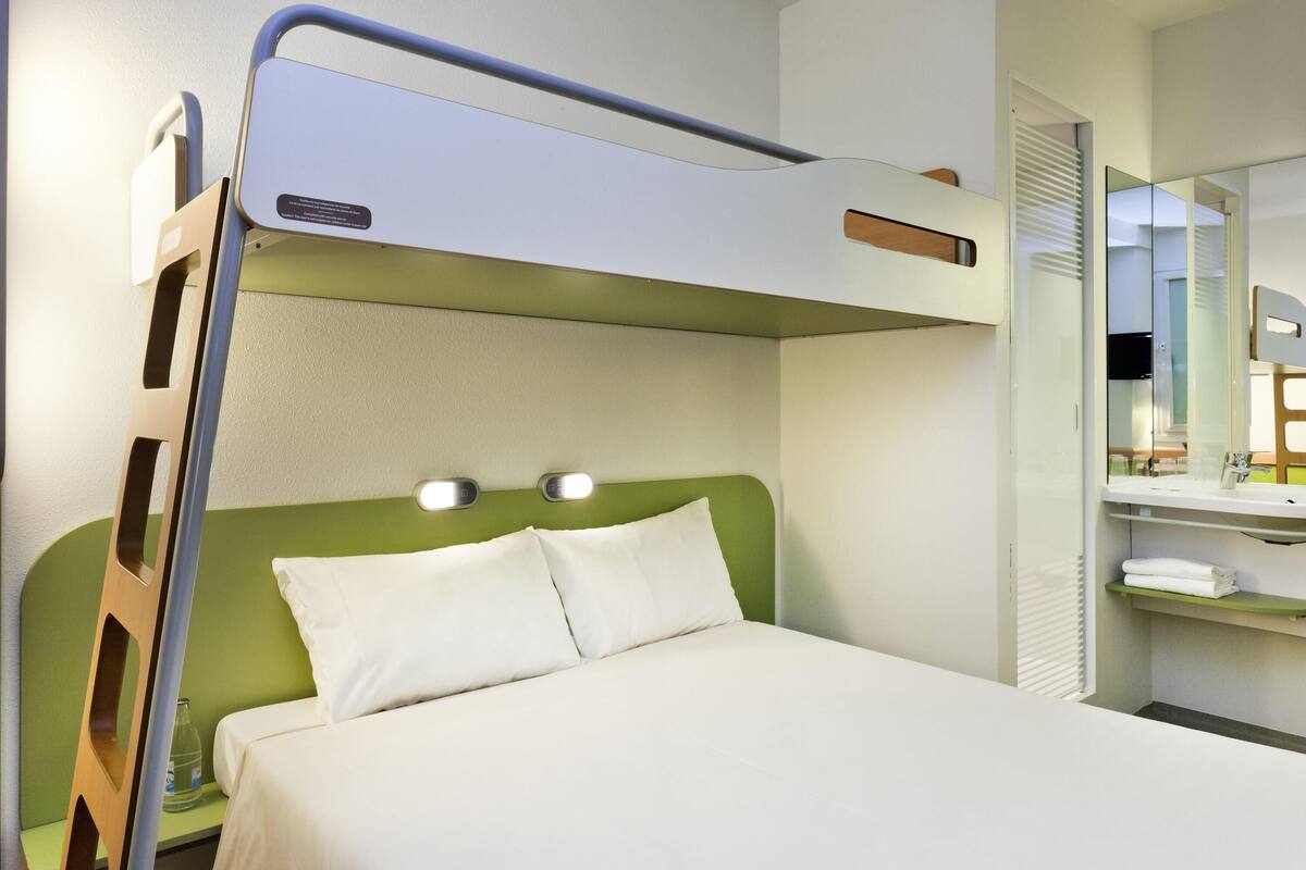 Uma cama beliche com cabeceira verde e roupa de cama branca, uma mesa de cabeceira com uma garrafa e um espelho na parede.