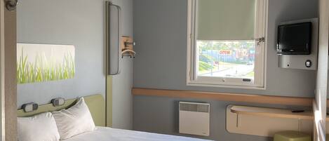 Une chambre d’hôtel avec un lit, un petit tabouret, un téléviseur fixé au mur et une fenêtre donnant sur l’extérieur.