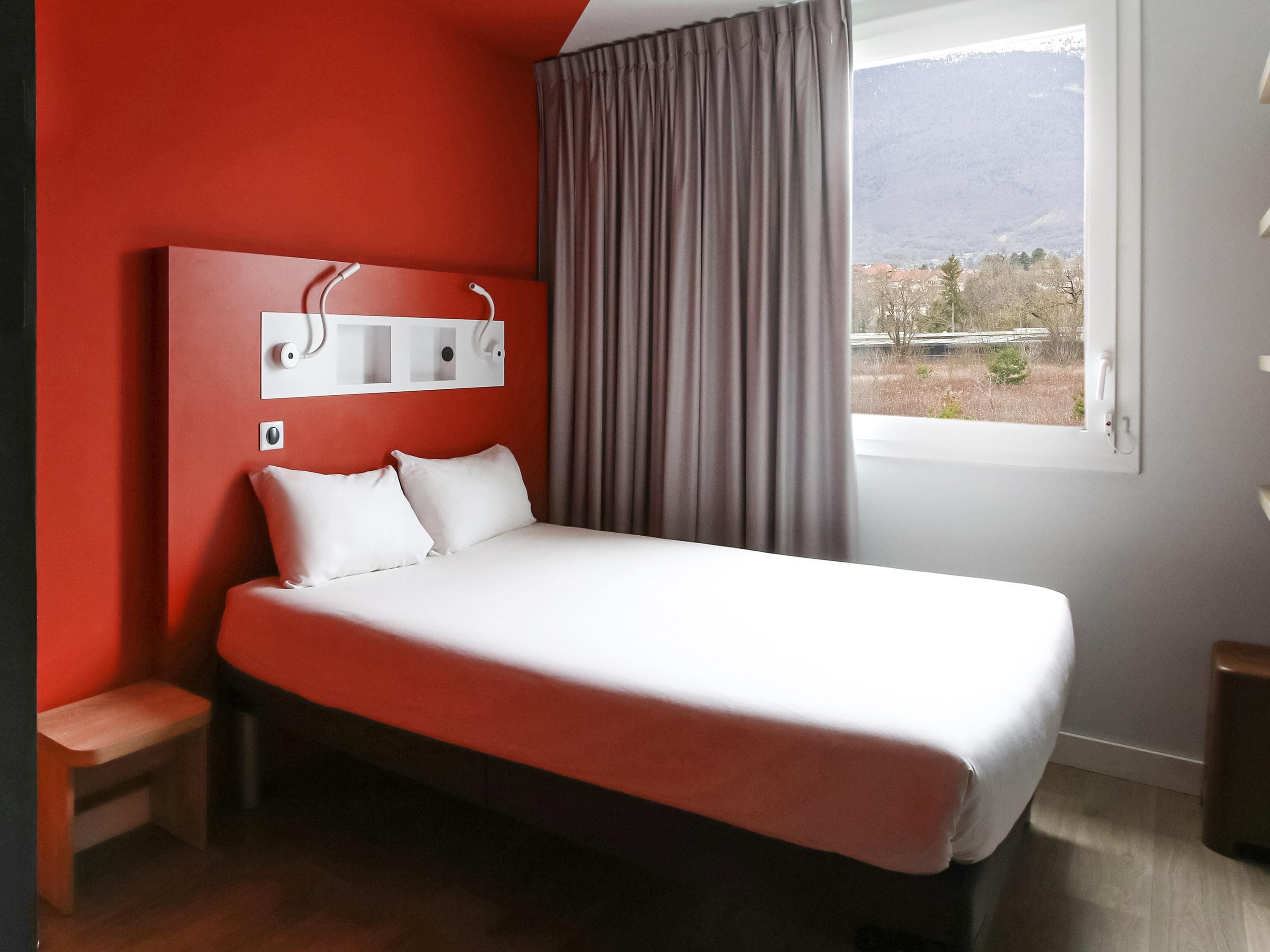 Ein Schlafzimmer mit Bett, Nachttisch, Fenster und Blick auf Berge, sowie einer roten Wand.