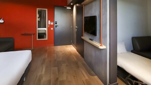 A hotel room with a bed, a desk, a chair, a TV mounted on the wall, and a bathroom with a mirror and sink. - Ibis Budget Saint Genis Pouilly Geneva (Saint-Genis-Pouilly)
