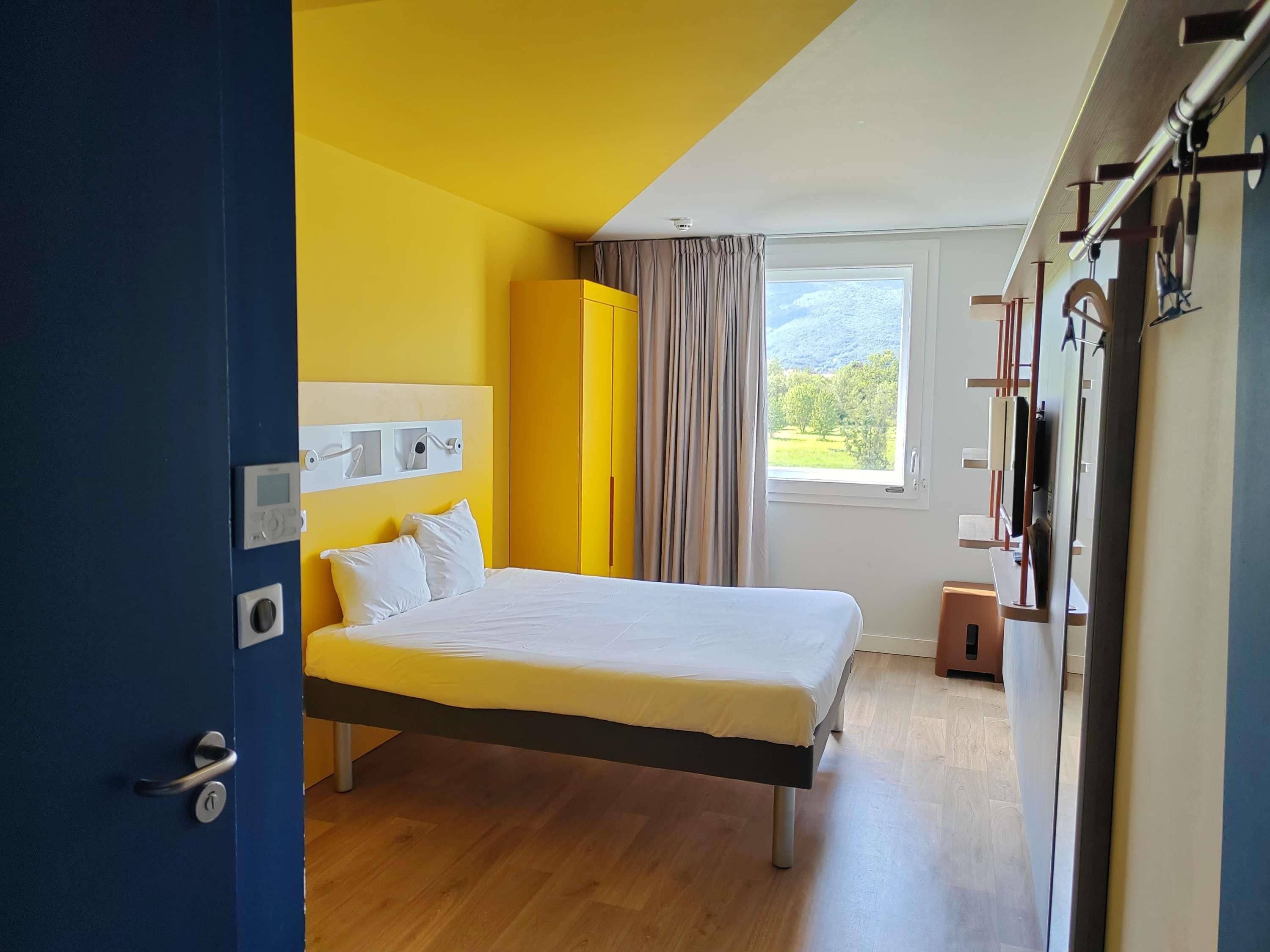 A modern hotel room with a bed, a window with a view, a wardrobe, and a door.