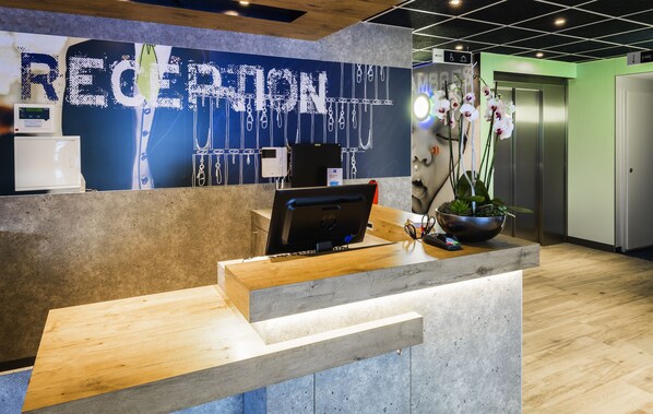 A reception desk with a computer, a plant, and a wall with the word "RECEPTION" in large letters. - Ibis Budget Saint Genis Pouilly Geneva (Saint-Genis-Pouilly)