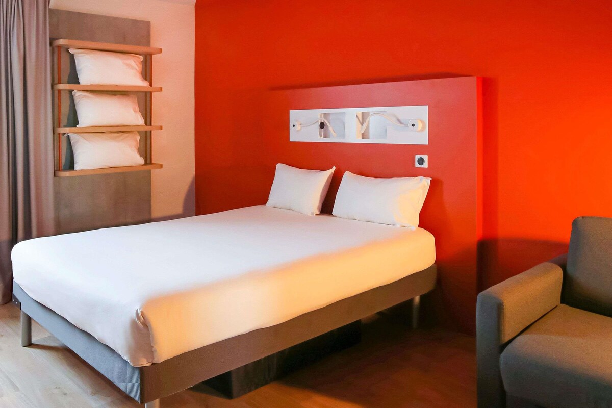 A hotel room with a bed, a chair, a shelf with pillows, and a red wall.