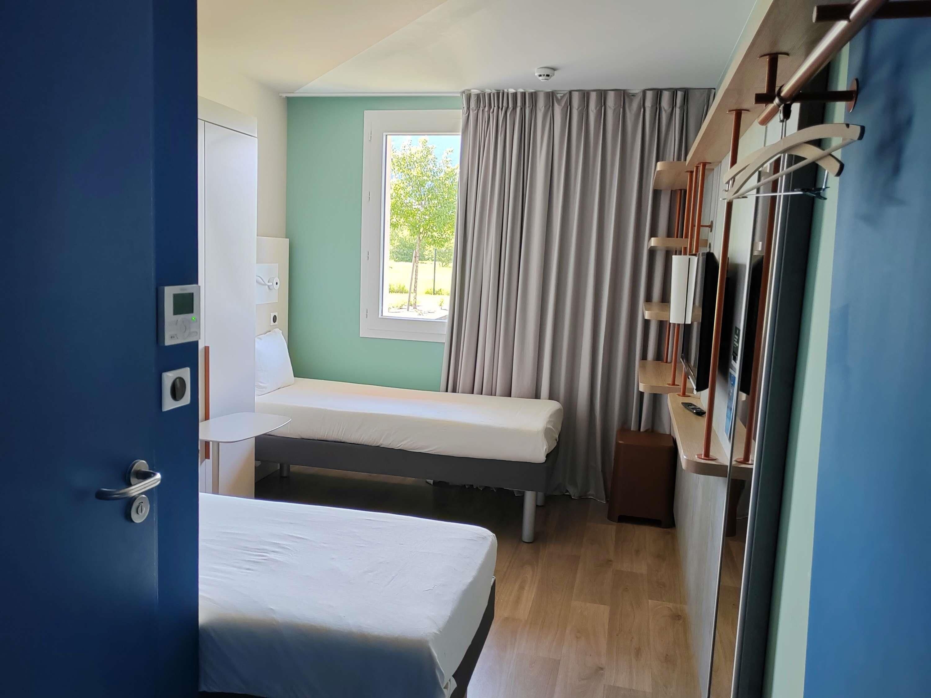 A room with a bed, a window with curtains, a bedside table, a wall-mounted TV, and a door with a handle.