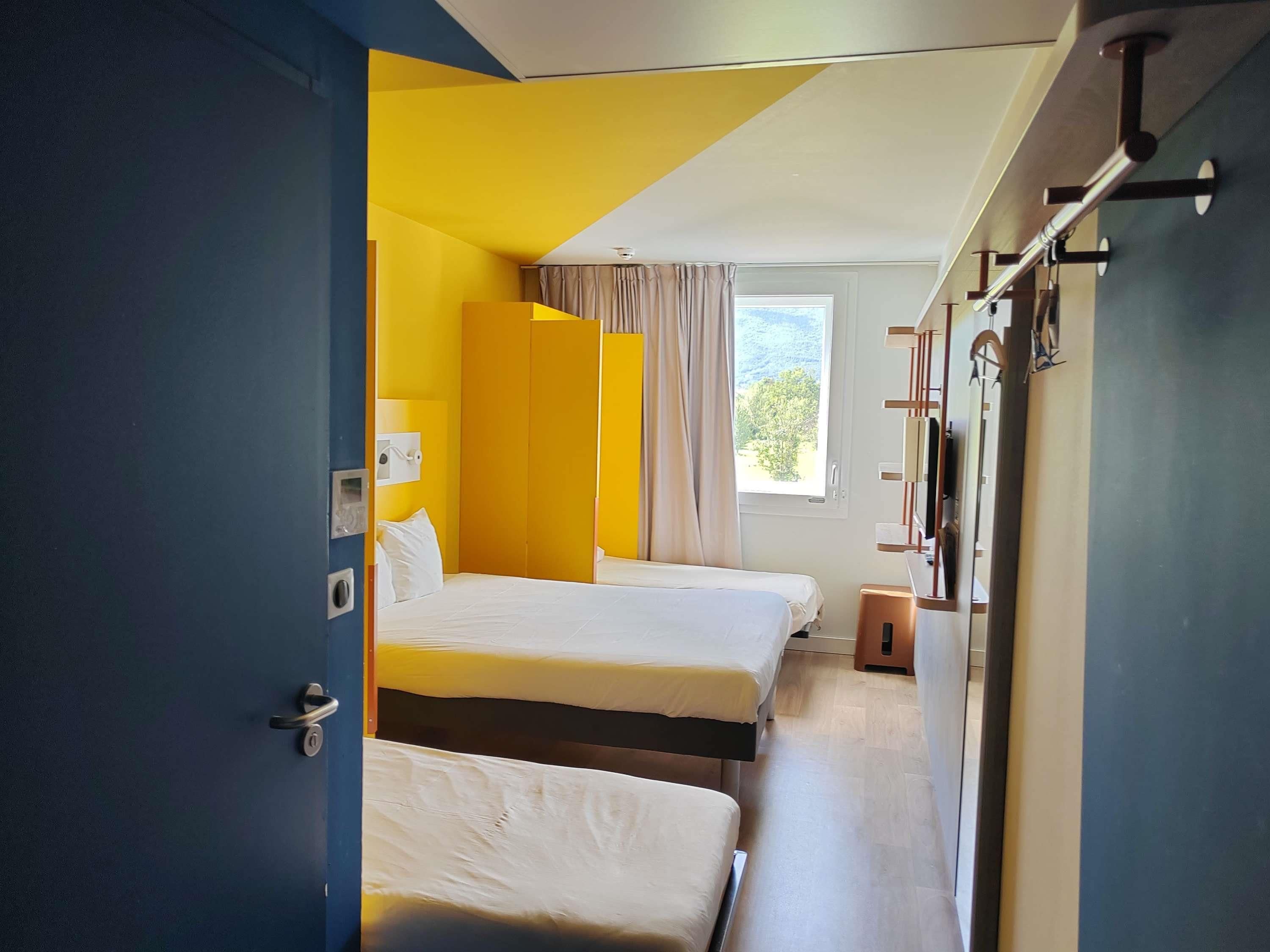 Ein Hotelzimmer mit zwei Betten, einem Fenster mit Vorhängen, einem Schreibtisch mit Stuhl und einem Kleiderschrank.