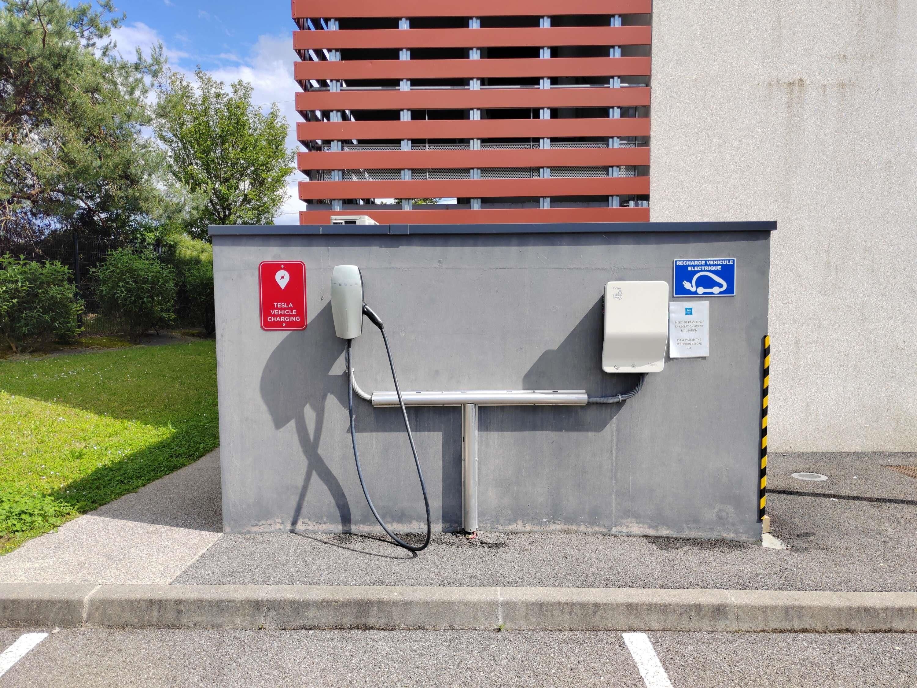 A parking area with a Tesla charging station, a wheelchair-accessible sign, and a building with a red facade in the background.