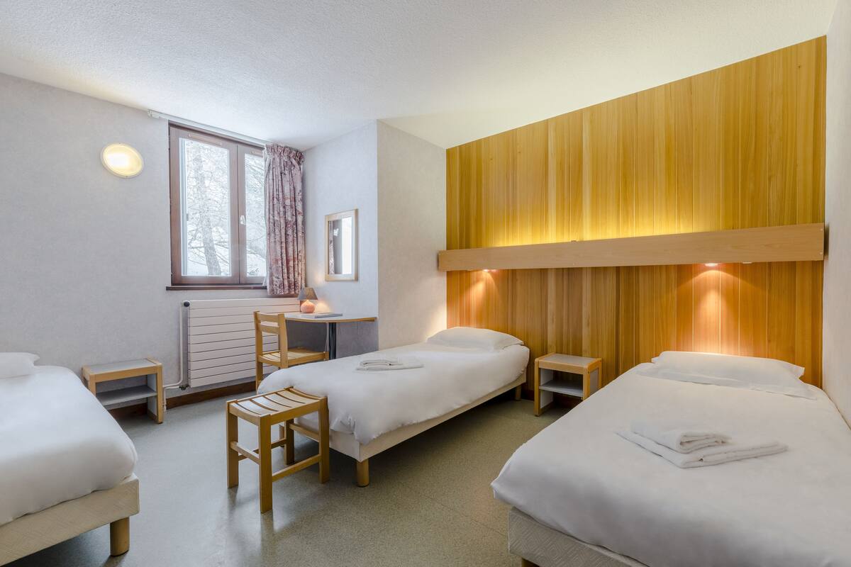 A hotel room with two single beds, a small desk, a chair, and a window with curtains.