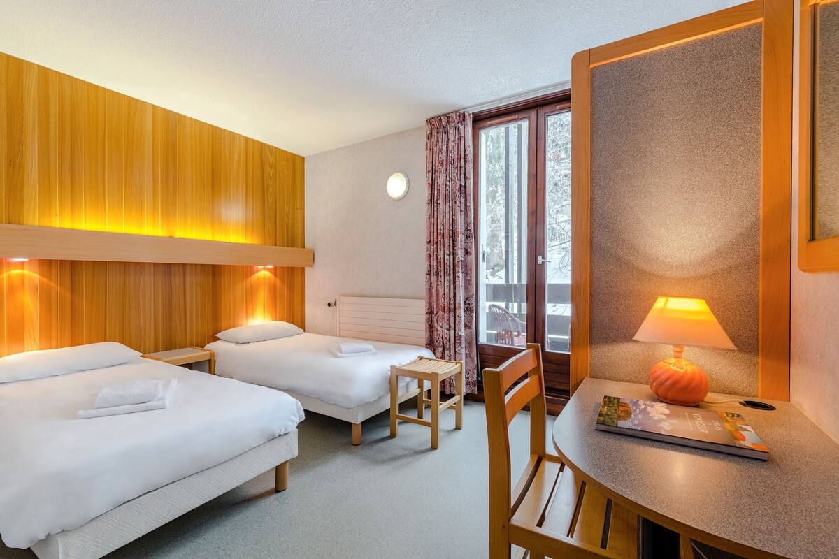 A hotel room with two beds, a small table, a chair, a lamp, and a view of the outdoors.