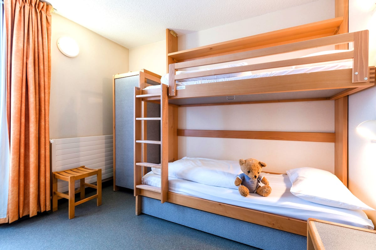 A bunk bed with a teddy bear on the lower bunk, a small wooden stool, and a window with curtains.