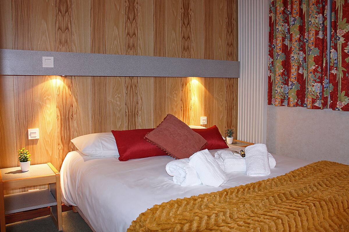 A hotel room with a wooden headboard, a bed with white linens, red and brown pillows, a yellow textured blanket, and a small potted plant on a nightstand.