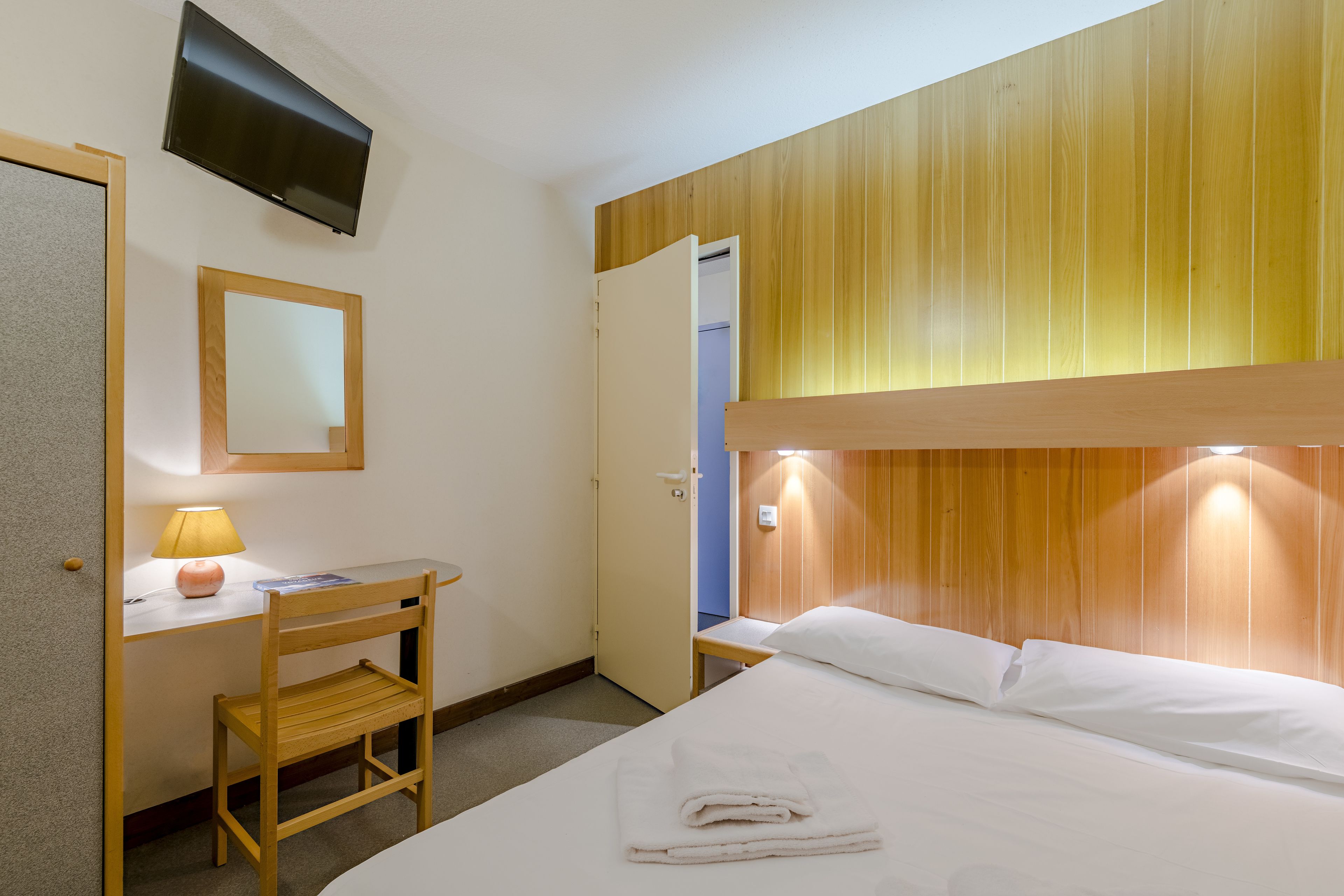 Hotelkamer met een bed, een bureau met stoel, een aan de muur bevestigde televisie en een houten paneeloplossing in het bovenste deel van de muur.