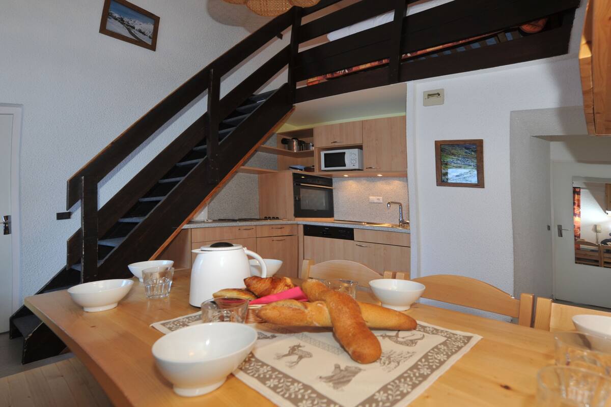 A dining area with a wooden table set for two, a kitchen in the background, and a staircase leading to an upper level.
