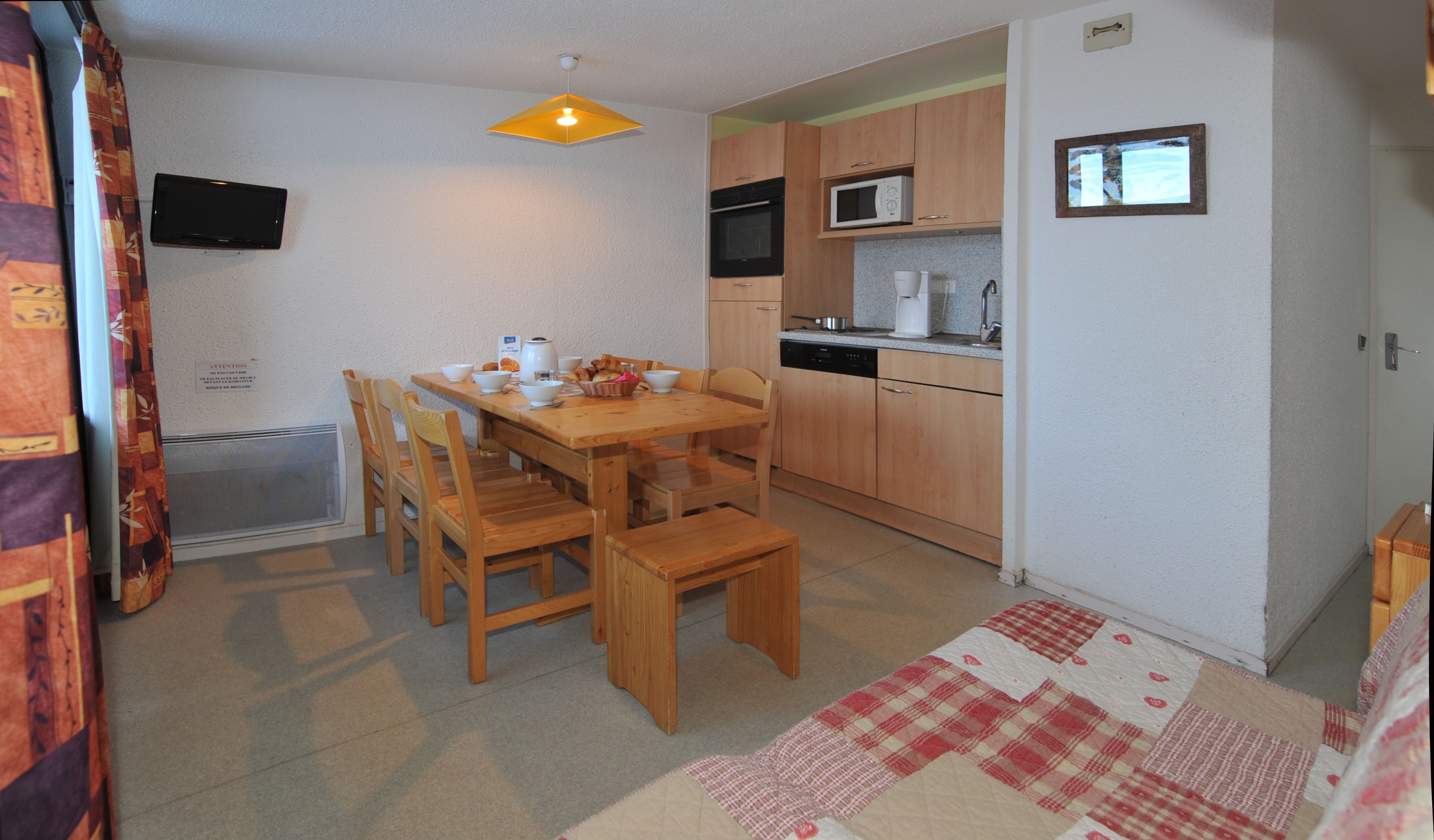 Cocina pequeña con armarios de madera, microondas y cafetera. Hay una mesa con sillas y una colcha en el suelo.