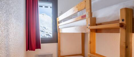 Een kleine, compacte kamer met stapelbedden, een houten bankje en een raam met een rode gordijn.
