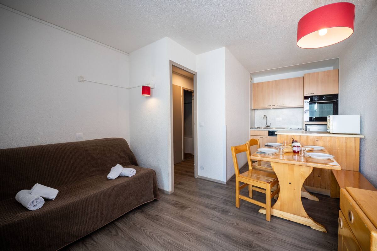 A compact living space with a brown sofa, a wooden dining table, and a kitchen area with wooden cabinets and appliances.
