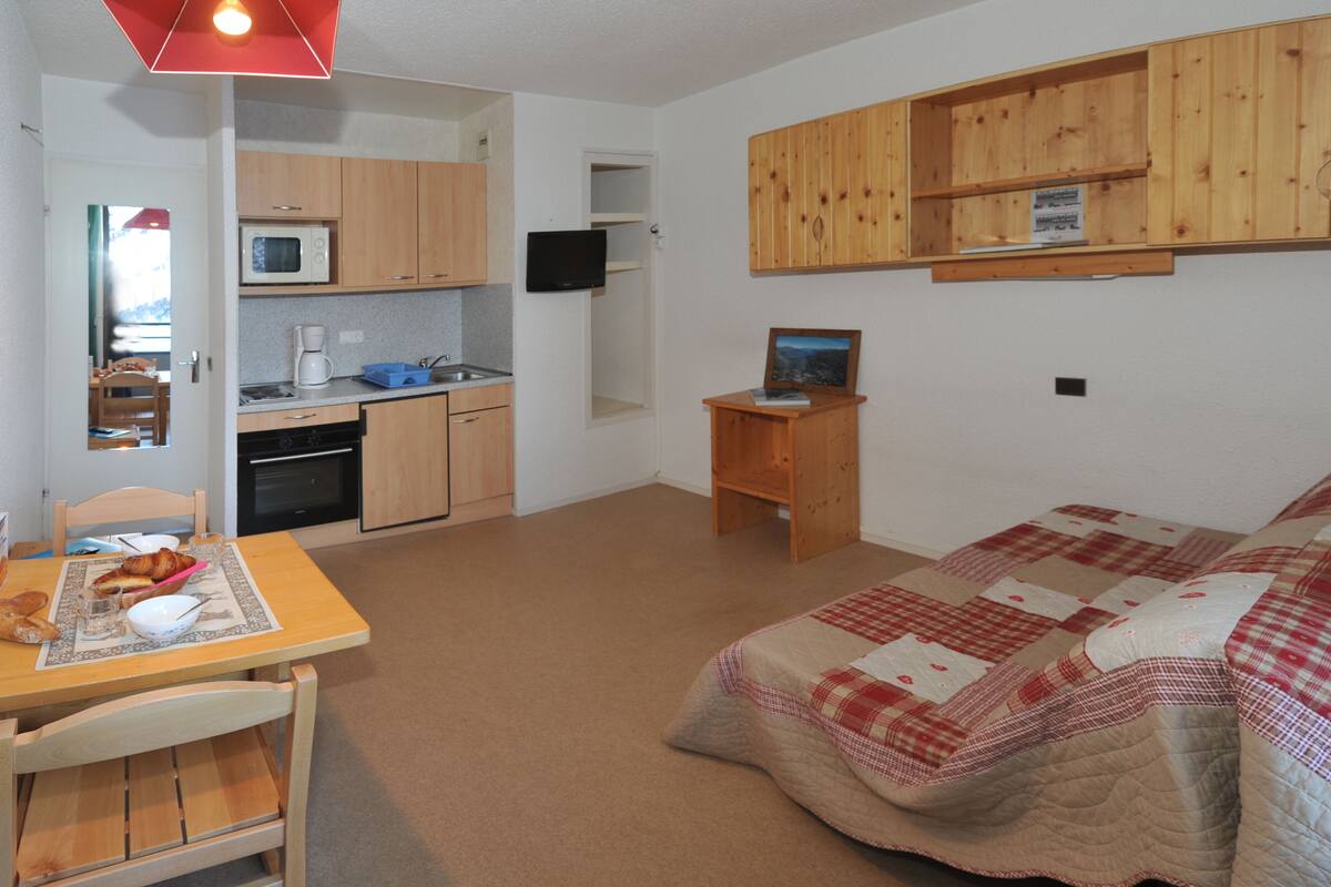 A compact room with a kitchenette, a bed with a checkered quilt, a small wooden table with breakfast items, and a television mounted on the wall.