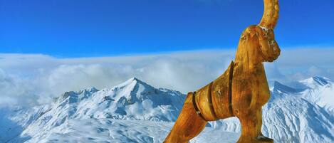 A sculpture of a creature with a curved horn, standing on a snowy mountain with a clear blue sky and snow-covered peaks in the background.